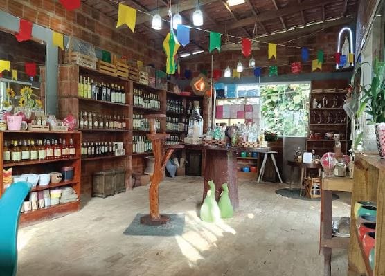 Interior da loja Flora Fogaça em São Benedito, com prateleiras de madeira expondo produtos regionais em garrafas de vidro, decoração de bandeirinhas coloridas e ambiente rústico iluminado por luz natural.